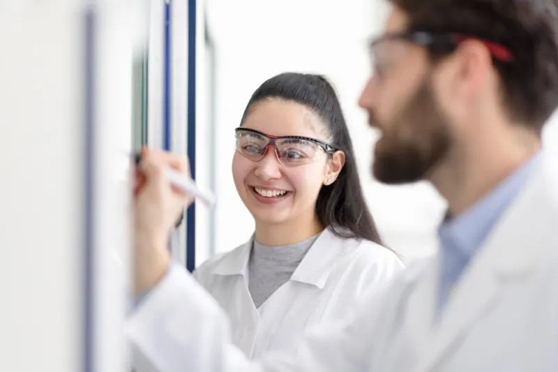 Lachende Mitarbeiterin mit Schutzbrille im Gespräch mit einem Kollegen im Labor